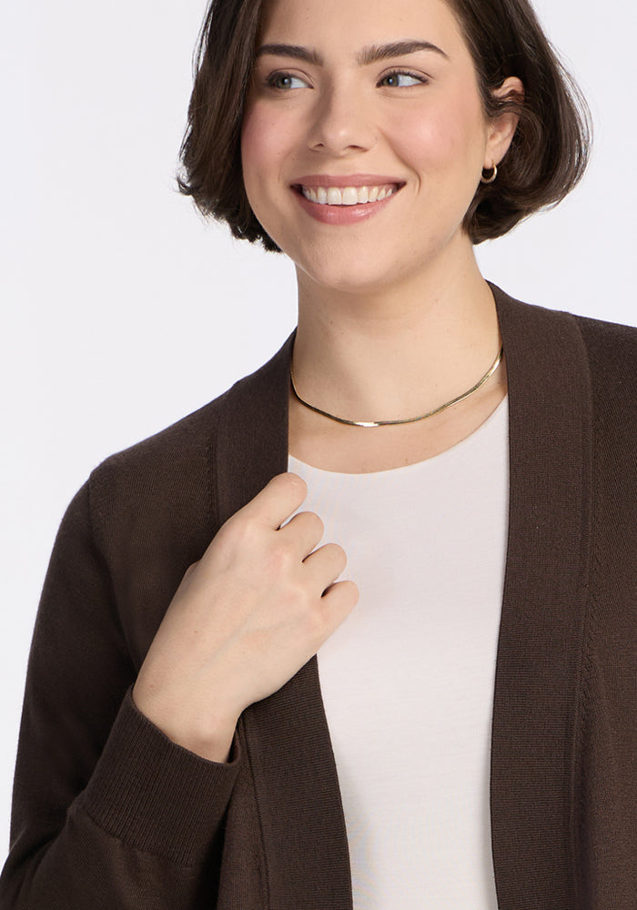 A woman with short brown hair smiles while wearing the Woolx Macie Sweater in Bracken over a white top, paired with a gold necklace and small hoop earrings, set against a plain light background.