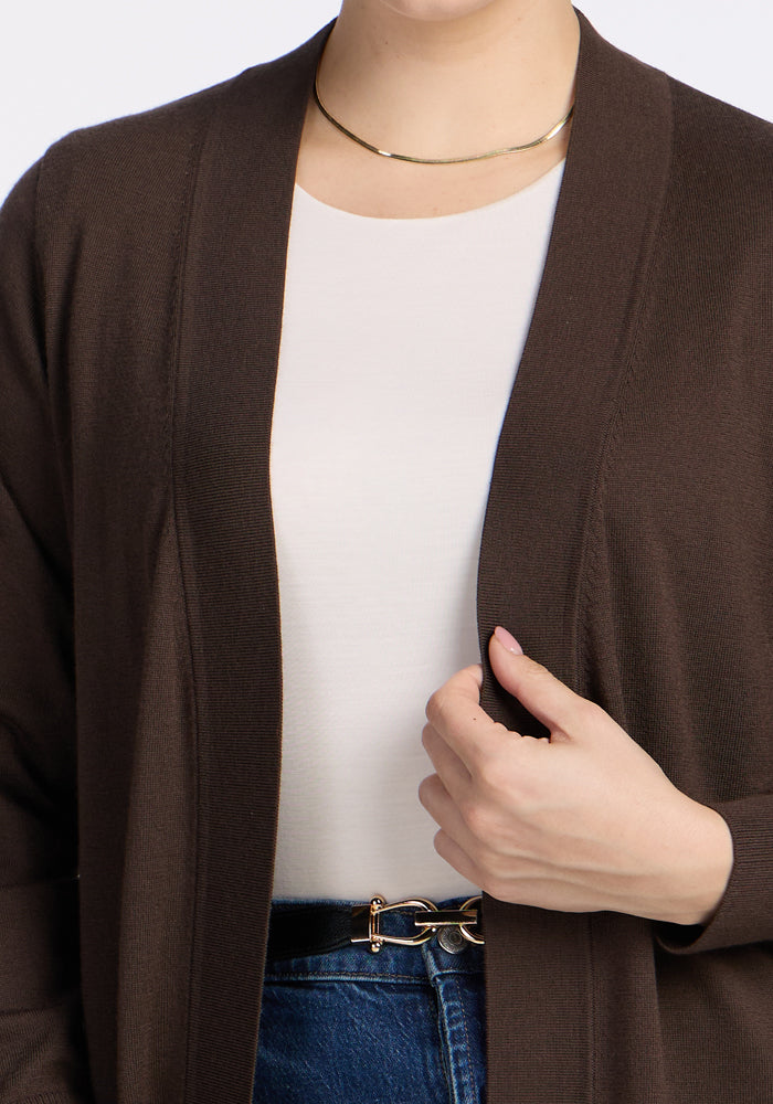 A person wears the Woolx Macie Sweater in Bracken over a white top, blue jeans with a gold-buckled belt, and a simple gold necklace, holding the edge of the cozy sweater with one hand.