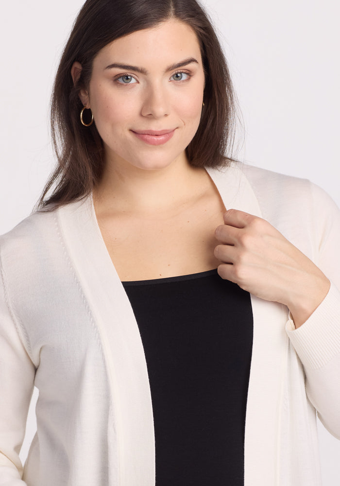 A person with long brown hair smiles at the camera, wearing the Woolx Macie Sweater in Dewdrop Pearl over a black top. They hold the sweater with one hand against a plain white background.