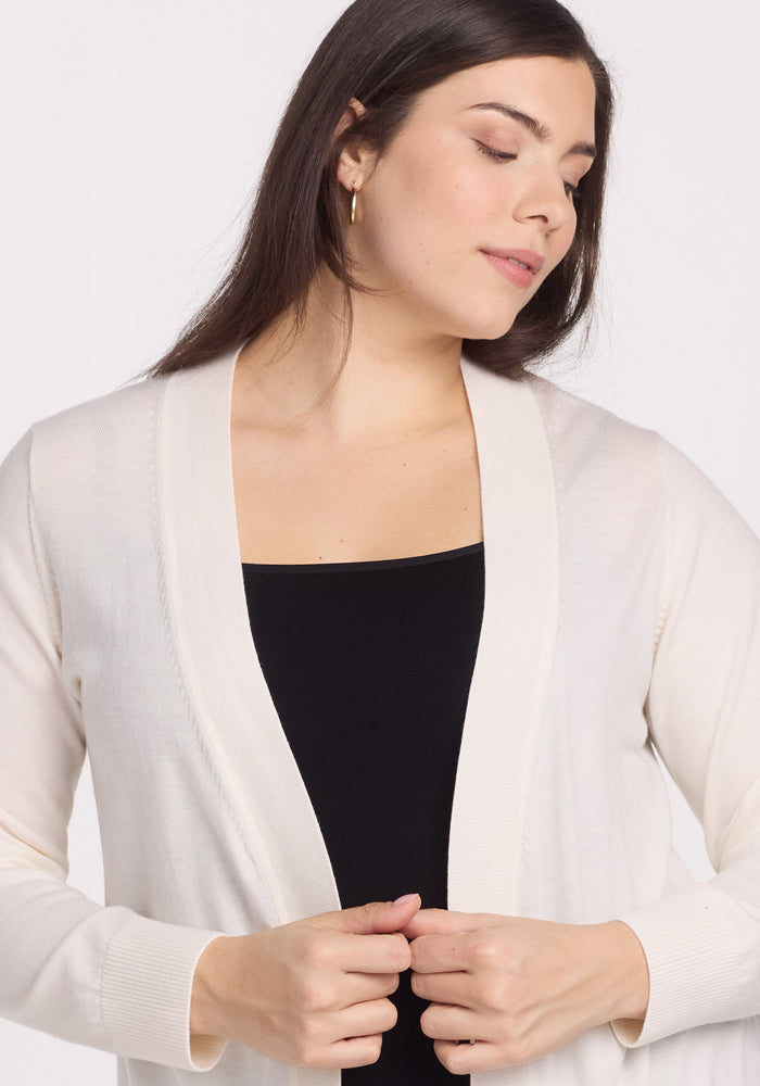A woman with long dark hair is pictured wearing a Woolx Macie Sweater - Dewdrop Pearl over a black top. She is looking down while holding the edges of the sweater against a plain white background.