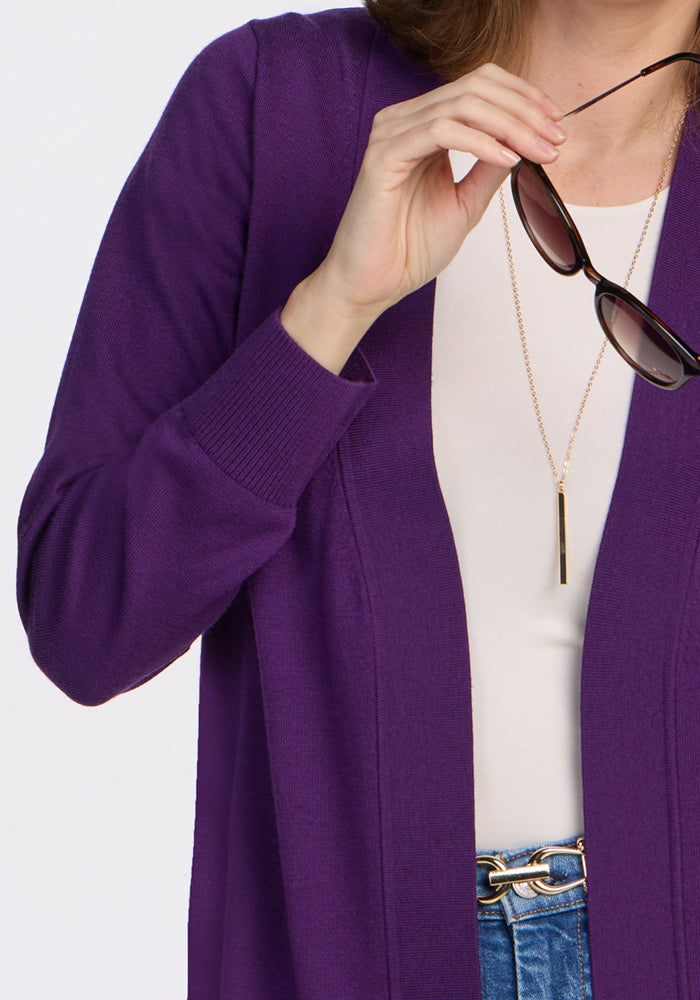 A woman models the Woolx Macie Sweater in Plum Crush over a white top and blue jeans, holding sunglasses near her face. She accessorizes with a gold vertical pendant necklace. The image is cropped at her shoulders and waist.