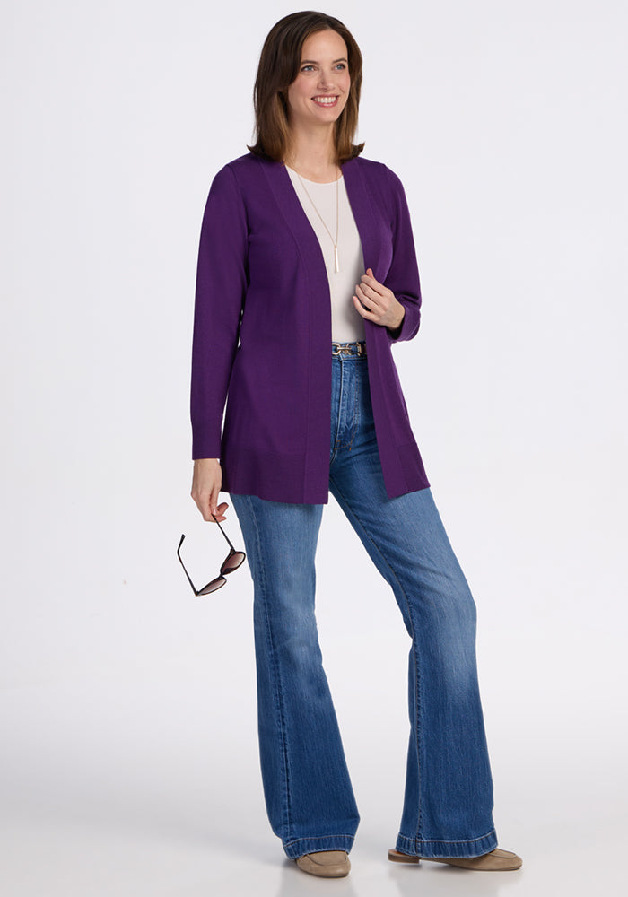 A woman smiles wearing the Woolx Macie Sweater in Plum Crush over a white top, blue flared jeans, taupe shoes, and a gold necklace. She has shoulder-length brown hair and holds sunglasses in her right hand.