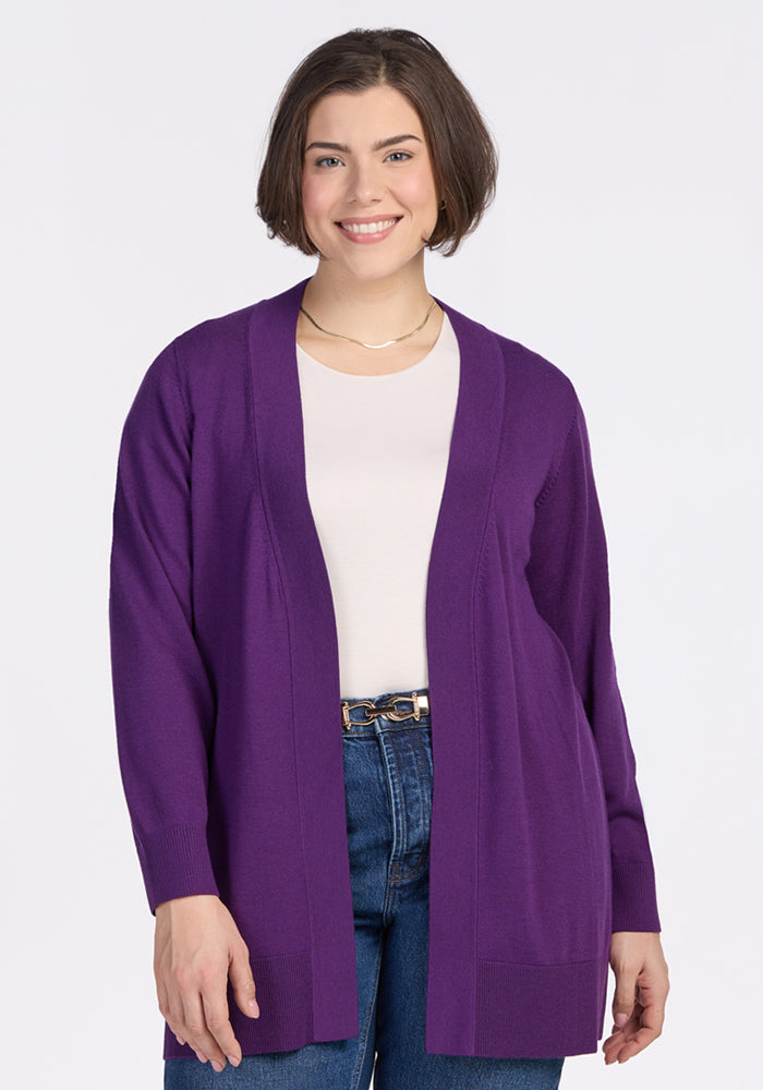 A woman with short brown hair smiles while wearing the Woolx Macie Sweater in Plum Crush over a white shirt and blue jeans, standing against a plain white background. | Juliana is 5'10", wearing a size XL
