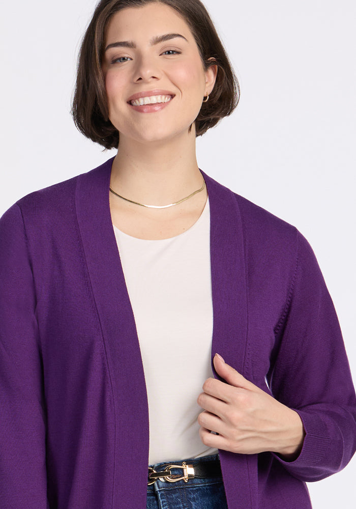 A smiling woman with short brown hair models the Woolx Macie Sweater in Plum Crush over a white top and blue jeans, accessorized with a gold necklace and hoop earrings, posing against a plain light background.