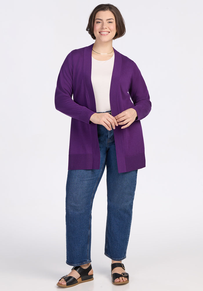 A woman with short brown hair smiles while facing forward. She wears the Woolx Macie Sweater in Plum Crush over a white top, paired with blue jeans and black sandals. The background is plain white.