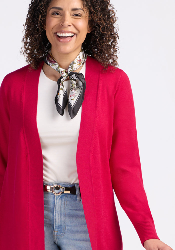 A woman with curly hair smiles in the Woolx Macie Sweater in Poppy Spark, styled open over a white top, blue jeans, a patterned scarf, and a belt with a circular buckle.