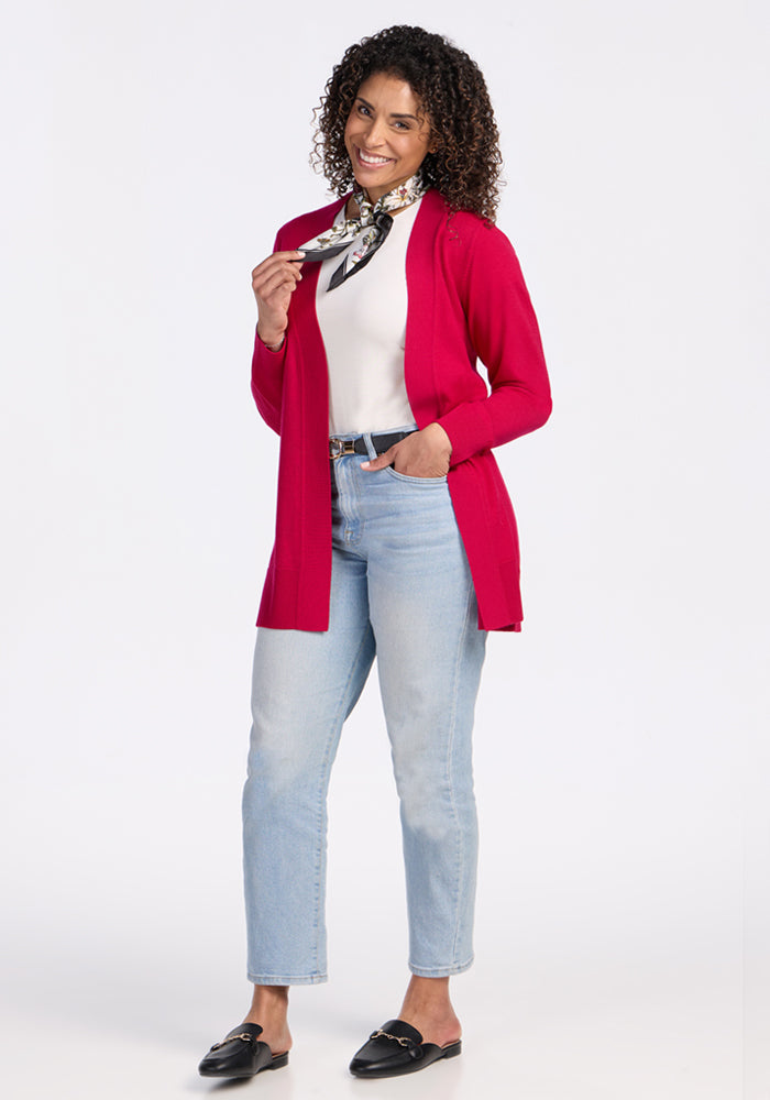 A smiling woman with curly hair models the Woolx Macie Sweater in Poppy Spark, paired with a white top, light blue jeans, black loafers, and a black-and-white patterned scarf. One hand is in her pocket and the other holds her scarf.