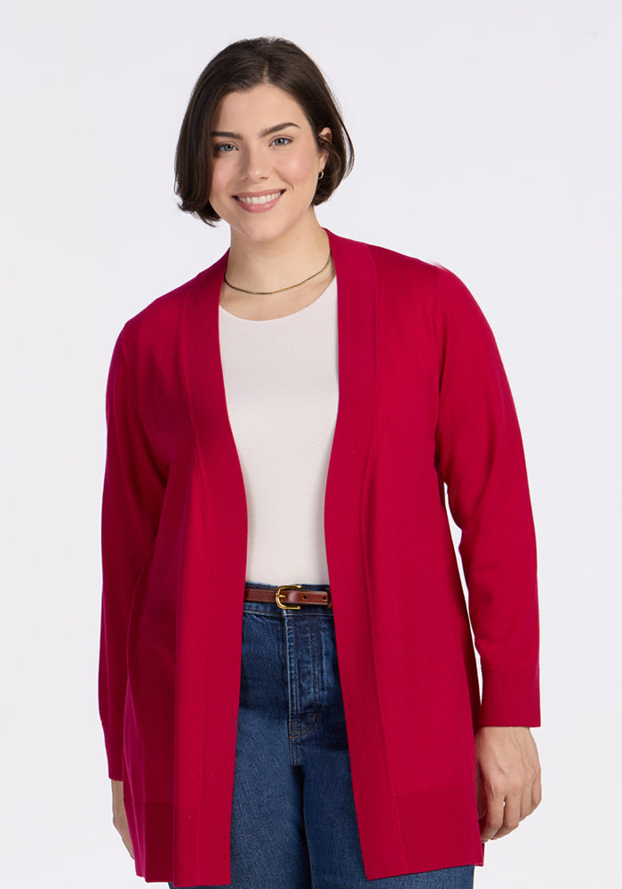 A person with short dark hair smiles while wearing the Woolx Macie Sweater in Poppy Spark over a white top and blue jeans, standing against a plain white background. | Juliana is 5'10", wearing a size XL