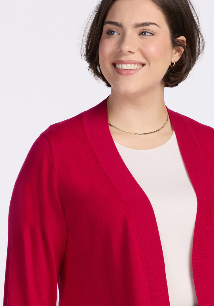 A woman with short brown hair is smiling while wearing the Woolx Macie Sweater in Poppy Spark over a light top and a thin gold necklace, set against a plain, light background.