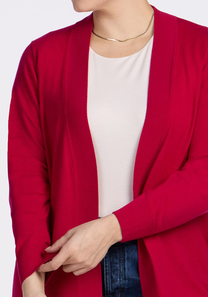 A person adjusts the sleeve of the Woolx Macie Sweater in Poppy Spark, worn open over a white top and blue jeans with a gold necklace. The cozy sweater is shown cropped to display only the upper torso and hands.
