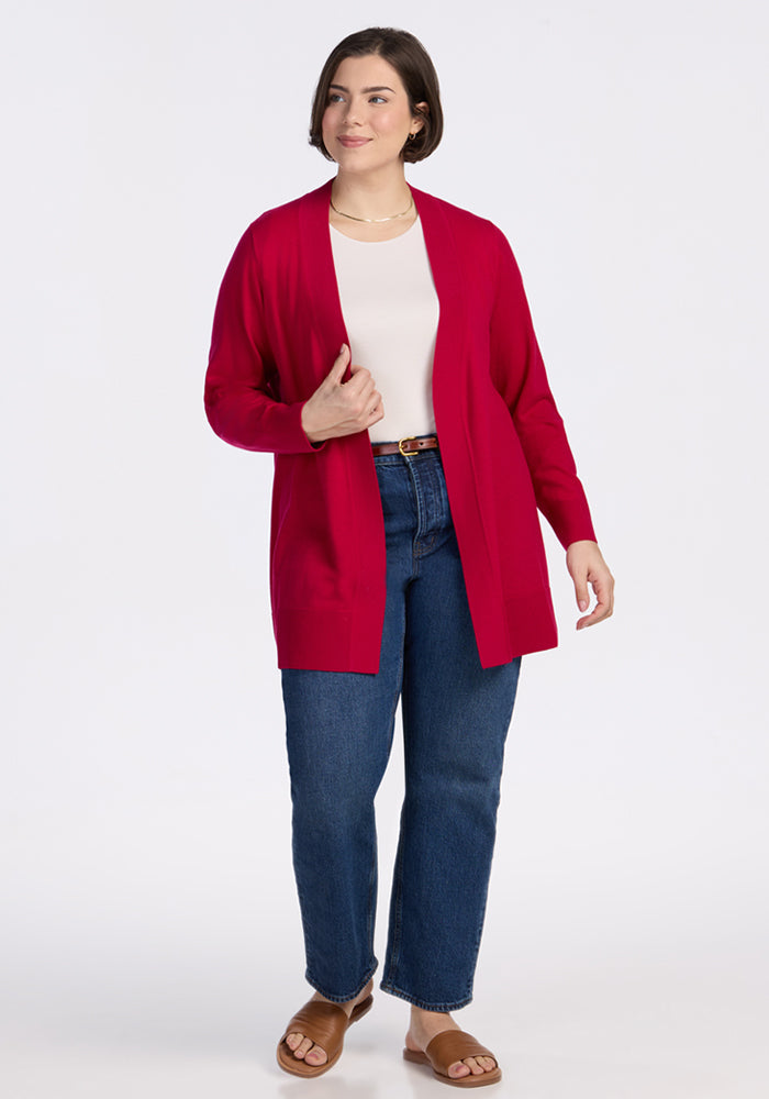 A woman with short brown hair wears the Woolx Macie Sweater in Poppy Spark over a white top with blue jeans, a brown belt, and sandals, standing and smiling against a plain white background.