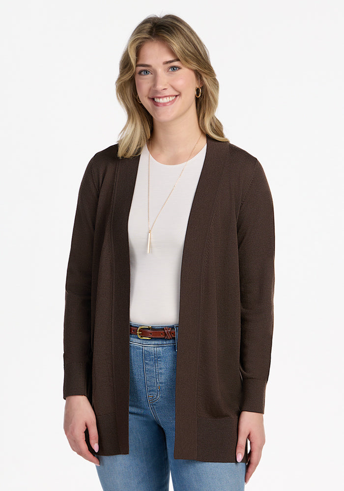 A blonde woman wearing the Woolx Macie Sweater - Bracken over a white shirt and blue jeans, smiling while standing against a plain white background. | Ariana is 5'11", wearing a size S