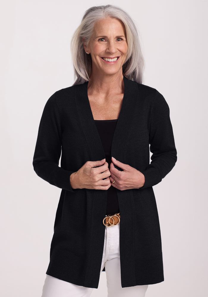 A smiling older woman with long gray hair models the Woolx Macie Sweater in Black over a matching top and white pants, standing against a light background. | Tam is 5’8.5”, wearing a size S
