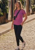 A smiling woman walks on a cobblestone street in a purple t-shirt, Woolx Madden Joggers - Black, white sneakers, and a black crossbody bag, with greenery and brick buildings in the background.