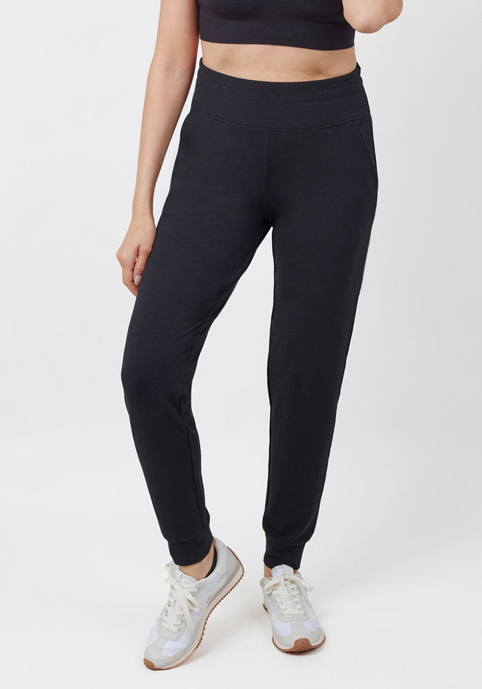 A person stands against a plain white background, shown from the shoulders down, wearing a black crop top, white sneakers, and Woolx Madden Joggers - Black.