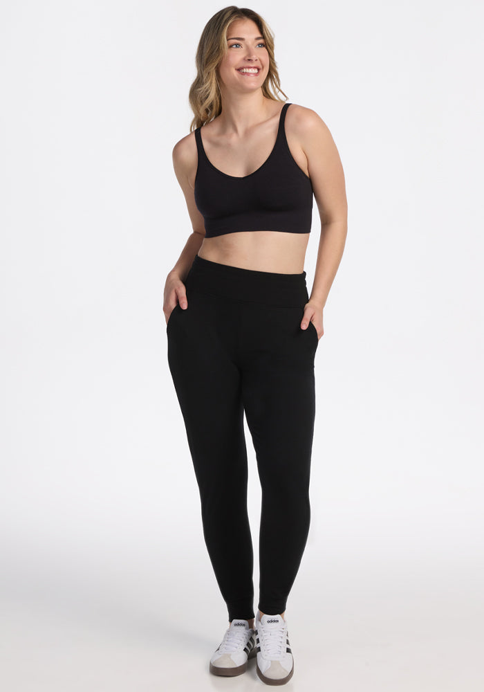 A woman smiles with her hands in the pockets of Woolx Madden Joggers - Black, paired with a black sports bra and white sneakers, against a plain white background. | Ariana is 5'11", wearing a size S