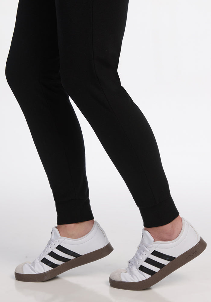 A person wears Woolx Madden Joggers in black with black and white sneakers featuring black stripes, standing on a white background. The image is cropped to show only the lower legs and feet.