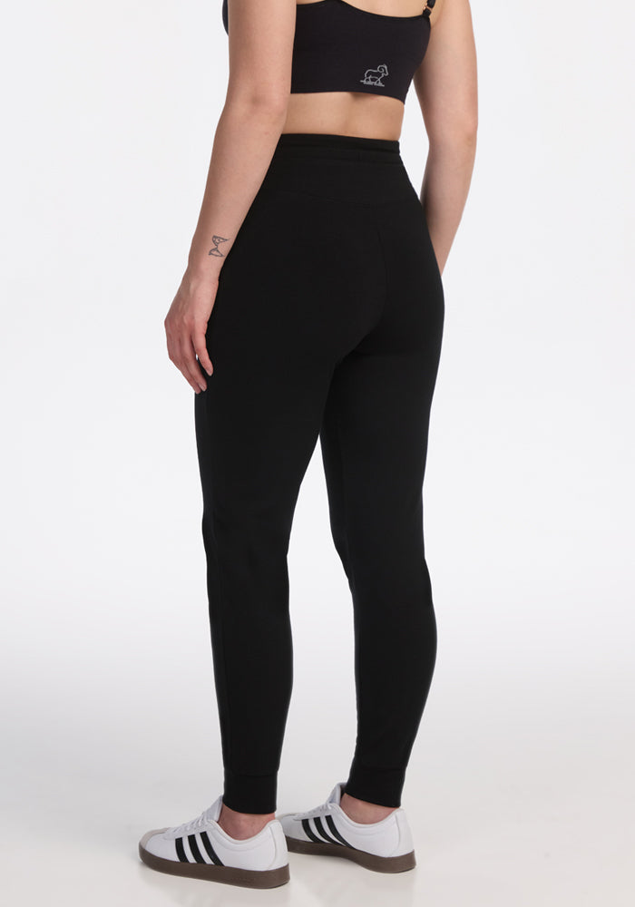 A person wearing a black sports bra, Woolx Madden Joggers - Black, and white sneakers with black stripes stands with their back to the camera against a white background.