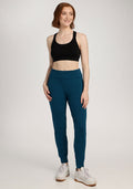 Woman in Madden Joggers - Majolica Blue by Woolx, black sports bra, and white sneakers standing and smiling.