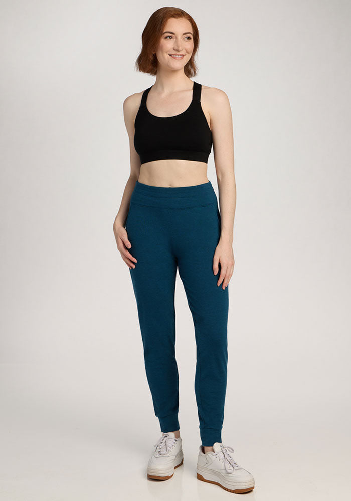 Woman in Madden Joggers - Majolica Blue by Woolx, black sports bra, and white sneakers standing and smiling.