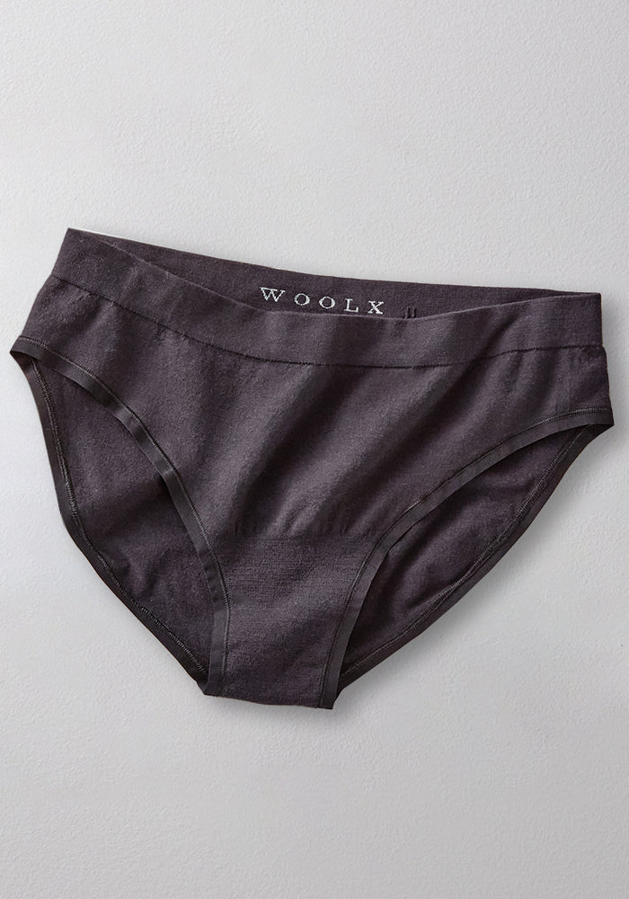 A pair of Woolx Maeve Bikini women's underwear in black, featuring a seamless design and branded waistband, laid flat on a light gray surface. Made from moisture-wicking merino wool for all-day comfort.