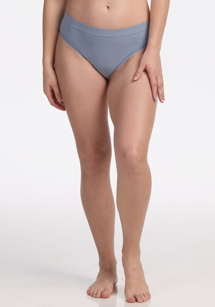 A person stands barefoot against a white background, wearing Woolx Maeve Bikini - Bluebell, moisture-wicking seamless underwear. Their arms rest at their sides, and their head is not visible in the image.