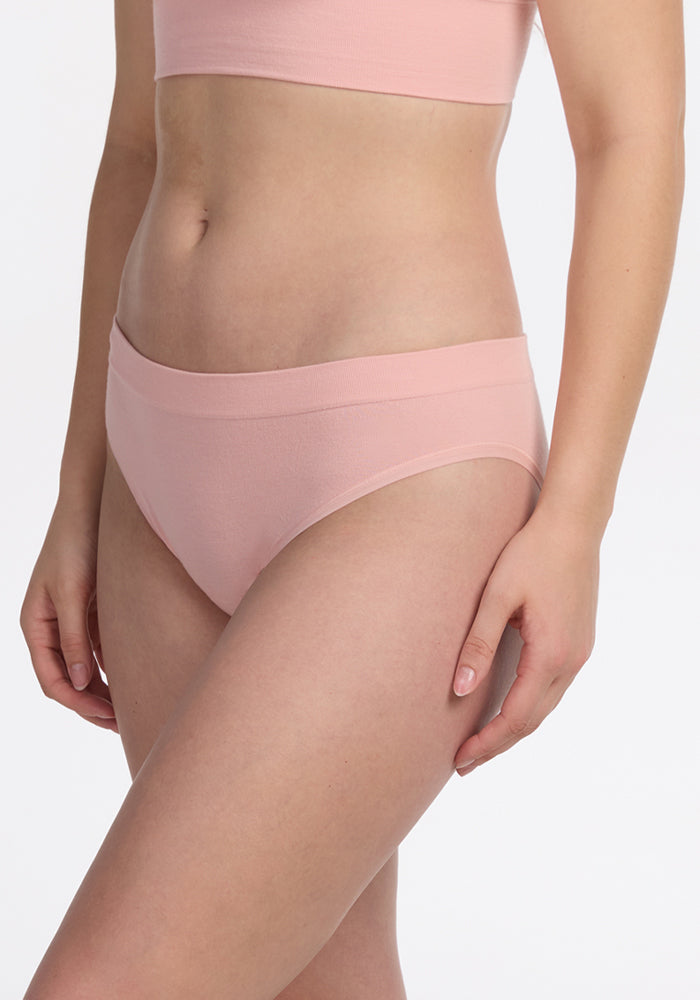 A person models the Woolx Maeve Bikini in Rosewater—a light pink seamless, moisture-wicking set—standing against a plain white background with one arm relaxed by their side.