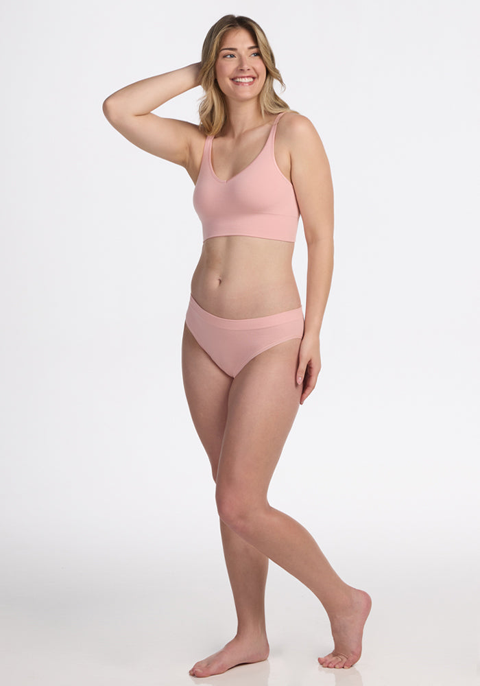 A woman stands barefoot against a white background, smiling with one hand on her head, wearing the Woolx Maeve Bikini in Rosewater—a seamless matching set that combines comfort and style.
