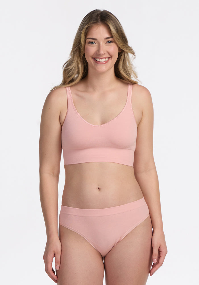 A woman with long, wavy blonde hair smiles at the camera, wearing the Woolx Maeve Bikini in Rosewater, standing against a plain white background.
