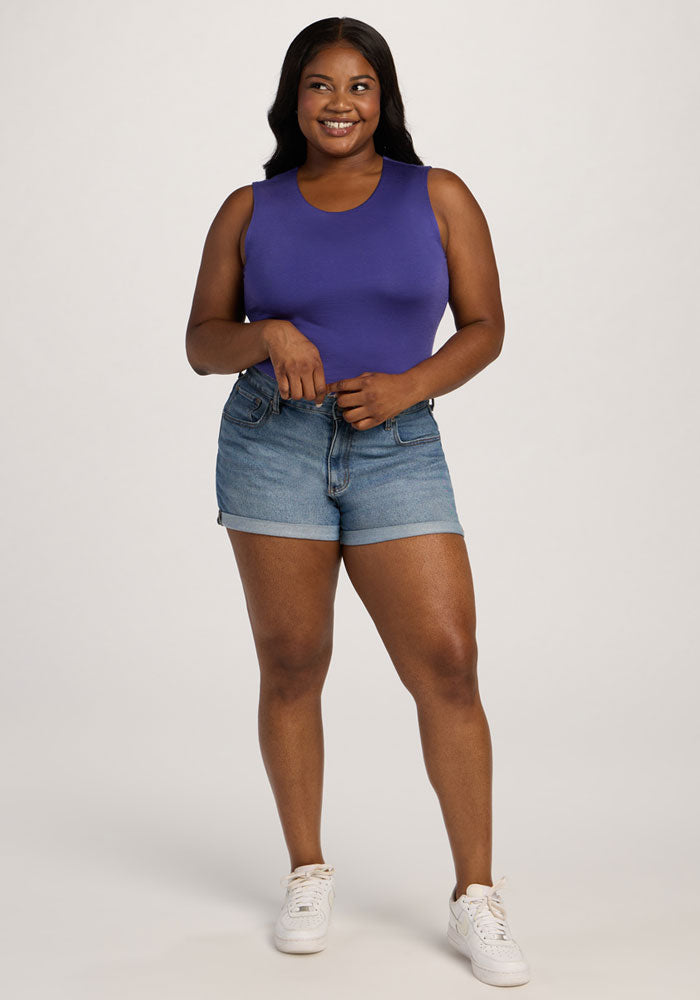 Woman smiling in a Woolx Maggie Bodysuit - Berry Indigo, denim shorts, and white sneakers against a plain background.