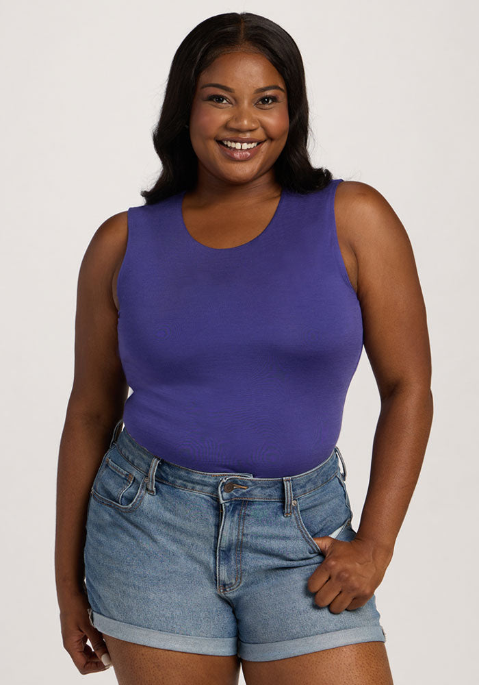 Smiling woman in the Woolx Maggie Bodysuit - Berry Indigo and denim shorts, standing against a plain light background. | Sapphire is 5’8”, wearing a size XL