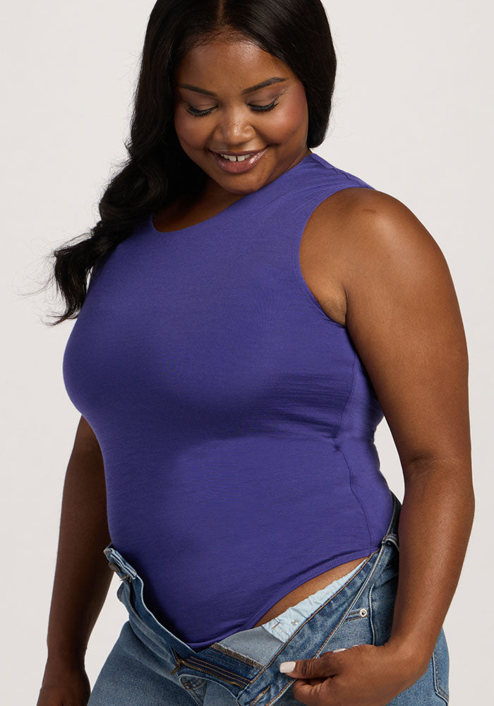 Woman smiles while looking down, wearing the Woolx Maggie Bodysuit - Berry Indigo with unbuttoned blue jeans.