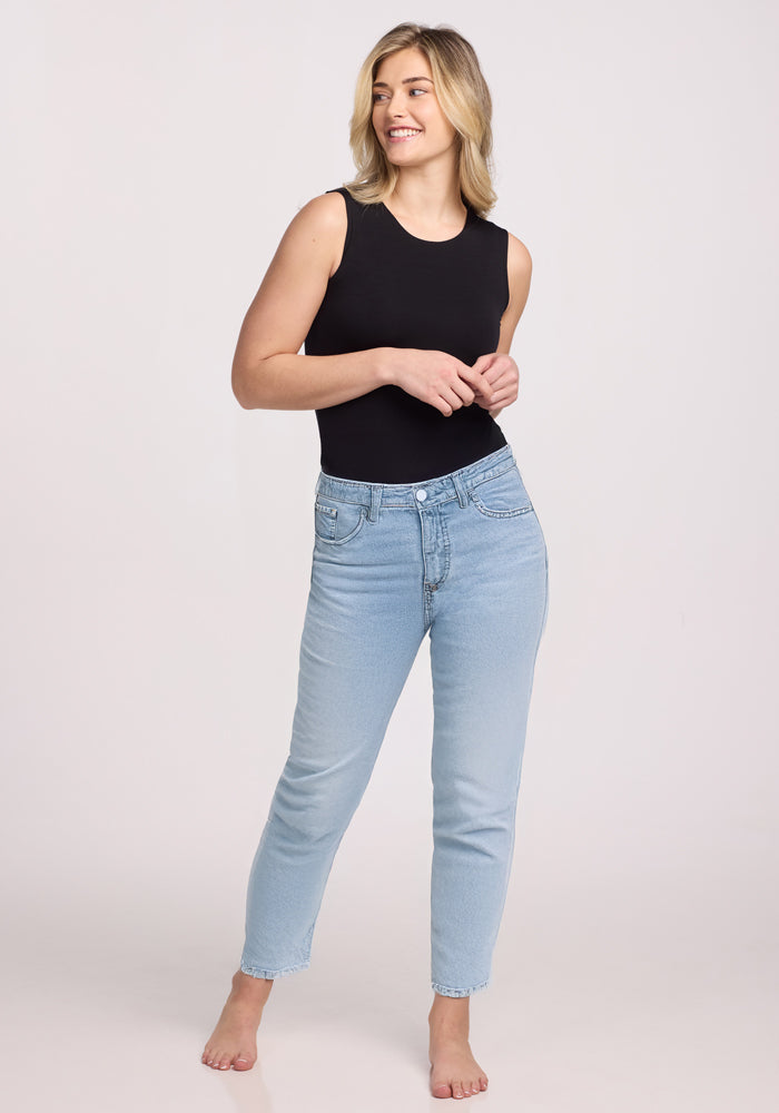 Standing barefoot against a plain background, they wear the Maggie Bodysuit - Black by Woolx and light blue jeans. With shoulder-length blonde hair, they smile confidently, facing slightly to the side in their temperature-regulating attire designed for all-day comfort.