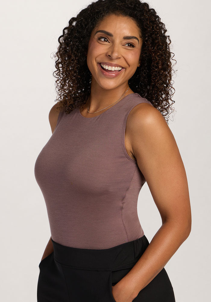 Smiling woman with curly hair wears the Woolx Maggie Bodysuit in Peppercorn and black pants against a plain background.