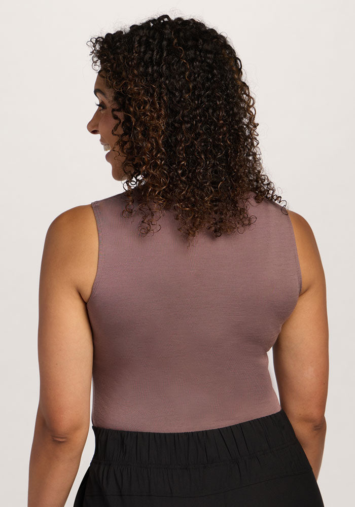 A woman with curly hair wears the Woolx Maggie Bodysuit in Peppercorn, seen from the back, with black pants.