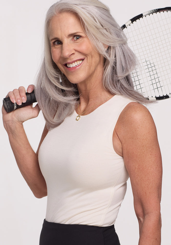 A smiling older woman with long gray hair holds a tennis racket, wearing the Woolx Maggie Bodysuit in Vanilla; her look is sleek and moisture-wicking against a plain light background.