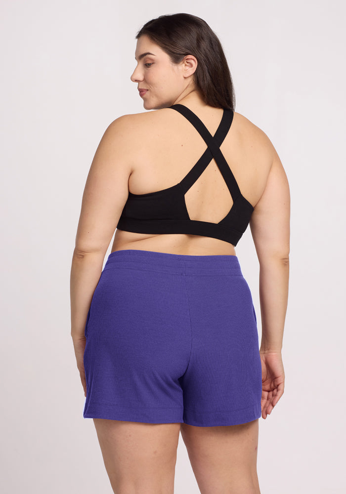 Woman stands with her back to the camera, wearing a black cross-back sports bra and Woolx Maisey Ribbed Shorts in Berry Indigo.