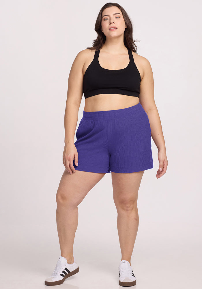 Woman in a black sports bra, Woolx Maisey Ribbed Shorts - Berry Indigo, and sneakers against a plain white background. | Juliana is 5'10", wearing a size XL