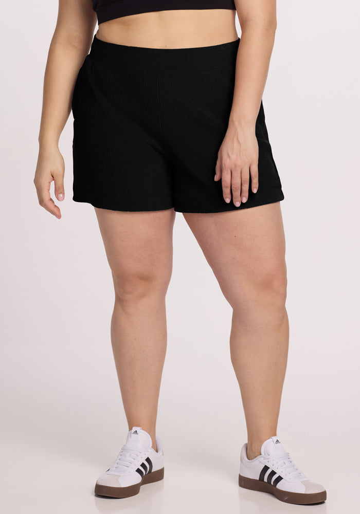 Standing against a plain background, a person models the lower half of their body wearing Woolx's Maisey Ribbed Shorts in black paired with white sneakers featuring black stripes. | Juliana is 5'10", wearing a size XL