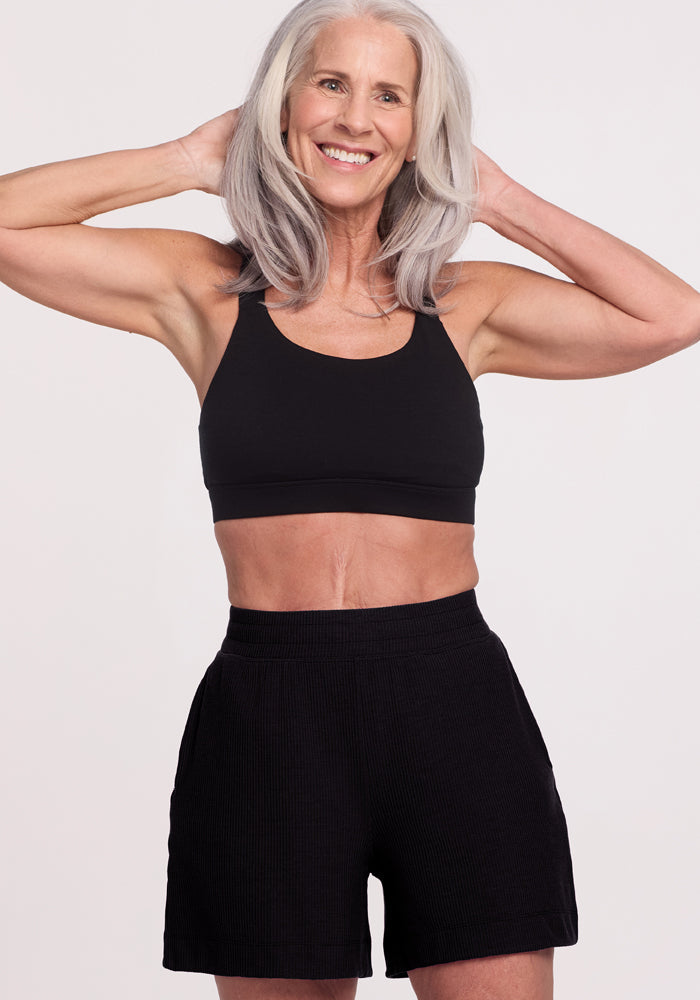 An older woman with long gray hair smiles, posing with her hands behind her head. She wears a black sports bra and Woolx's Maisey Ribbed Shorts - Black, set against a plain white background.