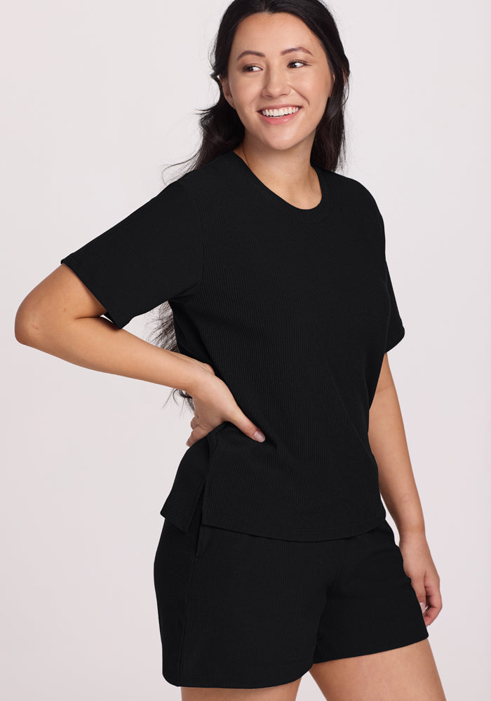With a relaxed and cheerful demeanor, a person with long dark hair poses against a plain light background in the Woolx Maisey Ribbed Shorts - Black, which has deep functional side pockets, and a black short-sleeve top.