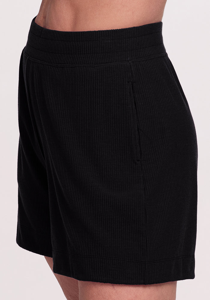 A person is shown from the waist down wearing Maisey Ribbed Shorts by Woolx, featuring a black ribbed texture, elastic waistband, side pockets, relaxed fit, and soft fabric.