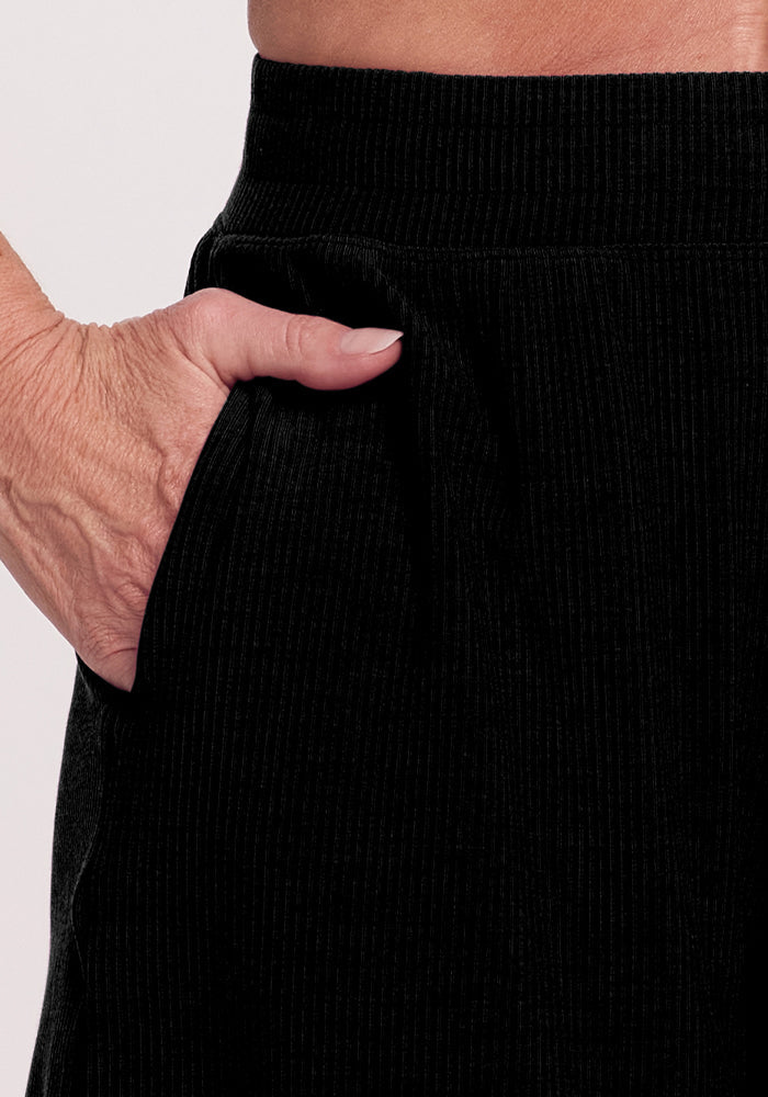 A close-up shows a hand in the pocket of black Maisey Ribbed Shorts by Woolx. The shorts have an elastic waistband, and the ribbed texture is visible against a plain, light-colored background.