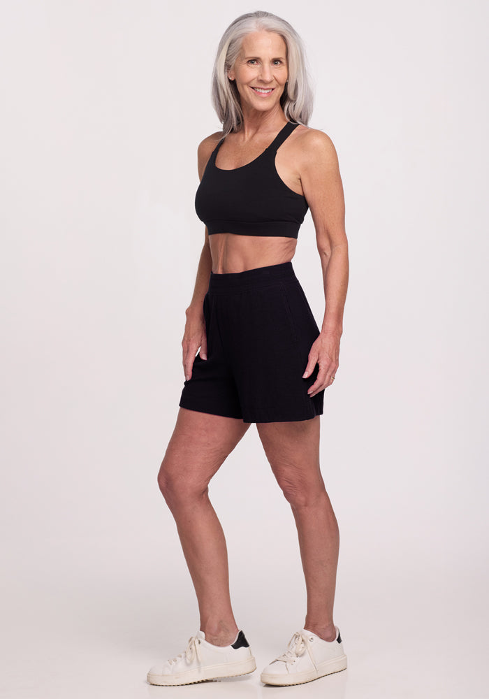 A smiling woman with long gray hair confidently poses in a black sports bra, Maisey Ribbed Shorts by Woolx, and white sneakers against a plain background.