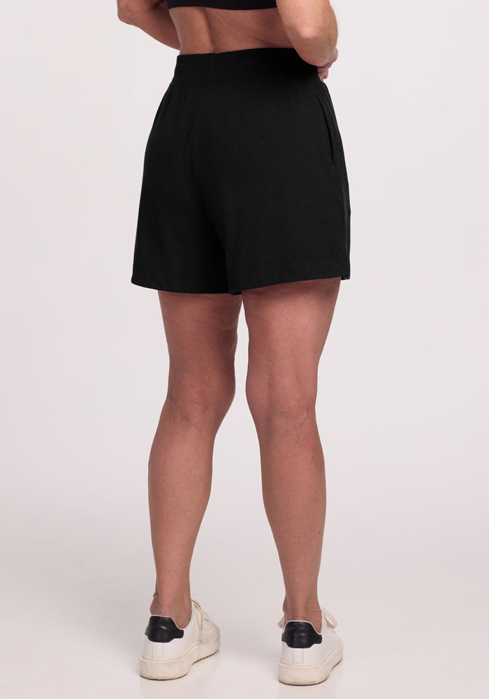 From behind, a person is seen wearing Woolx's Maisey Ribbed Shorts in black and white sneakers with black accents, standing against a plain white background.