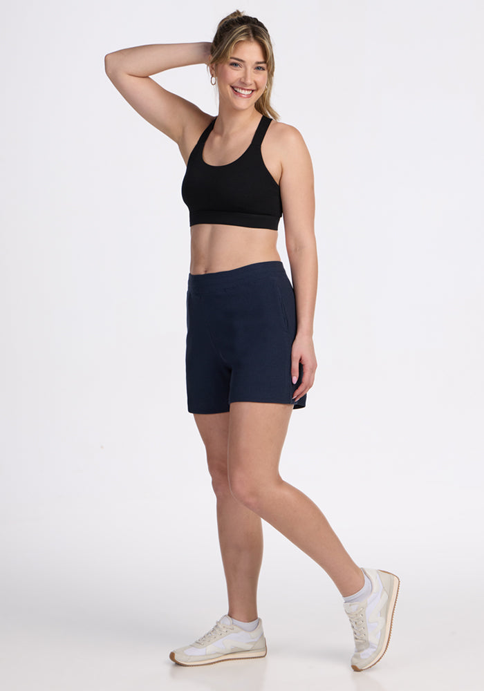 Smiling woman in a black sports bra, Woolx Maisey Ribbed Shorts in Deep Navy, and white sneakers poses with a hand up.