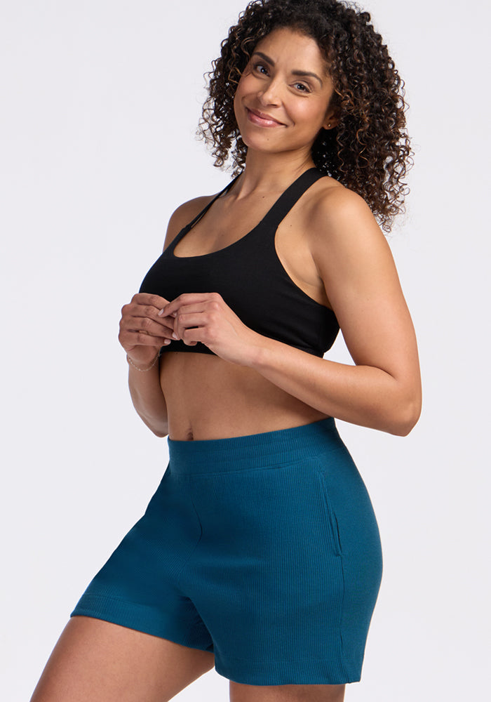 Smiling woman with curly hair wears Woolx Maisey Ribbed Shorts in Moroccan Blue and a black sports bra.