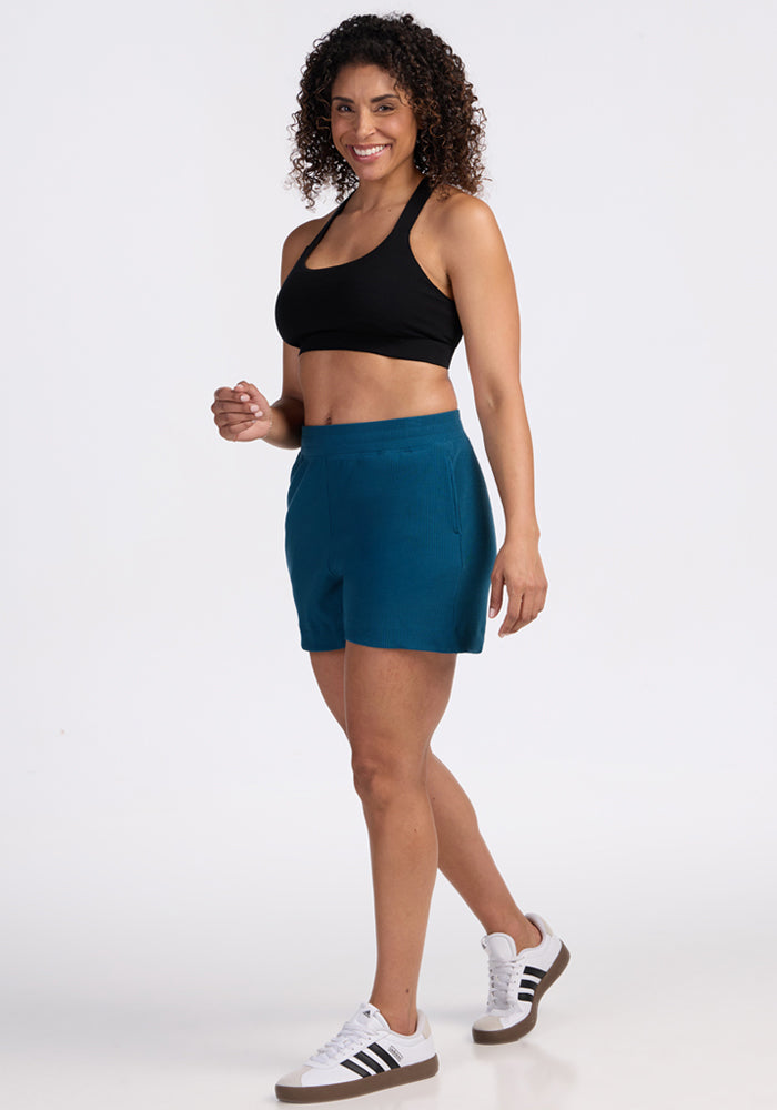 Woman in Woolx Maisey Ribbed Shorts in Moroccan Blue, black sports bra & white sneakers, smiling against a white background.