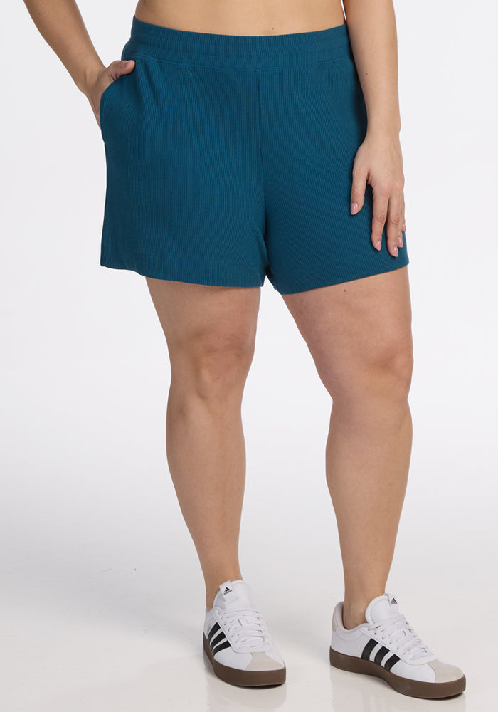 Person in Woolx Maisey Ribbed Shorts in Moroccan Blue and white sneakers, posing with a hand in pocket against white backdrop.