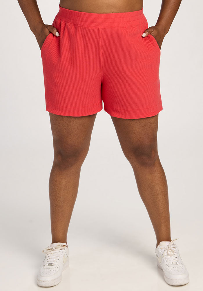 Model wearing Woolx Maisey Ribbed Shorts in Hibiscus with white sneakers, hands in pockets, on a plain background.